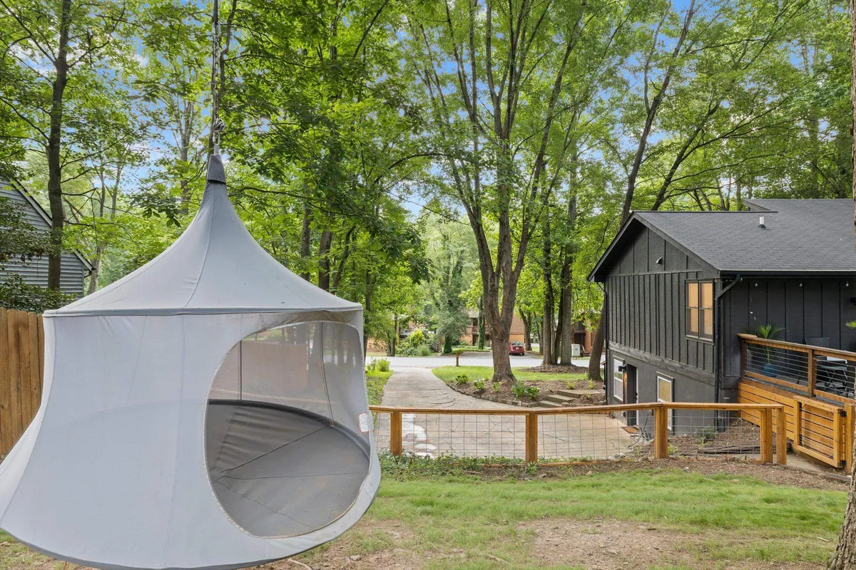 A peaceful backyard scene showcasing a swinging tent set up under trees, with the house in view and plenty of lawn space. There is open green space for play, the tent for relaxing or socializing, and a tranquil outdoor vibe perfect for family activities.