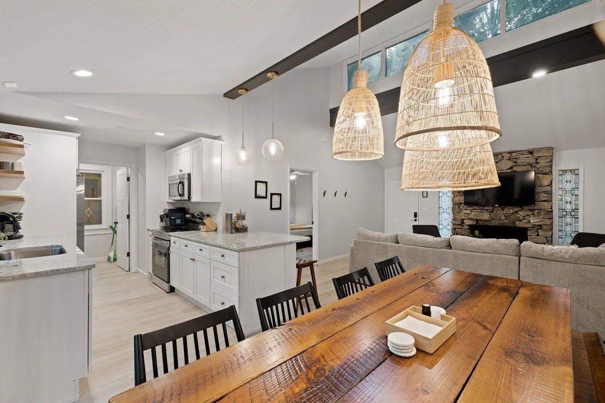An inviting open-plan space combining a kitchen island, dining table, warm pendant lighting, and a stone fireplace. The setting is styled for family meals, cooking together, and entertaining, with a visible flow between kitchen and dining areas.