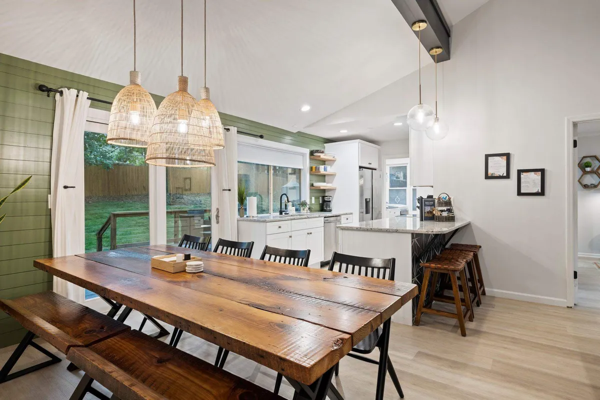 A spacious dining area featuring a large wooden table, woven pendant lights, and kitchen counter with barstools. Sliding glass doors lead directly to the backyard, suggesting easy access to outdoor dining or grilling on the deck.