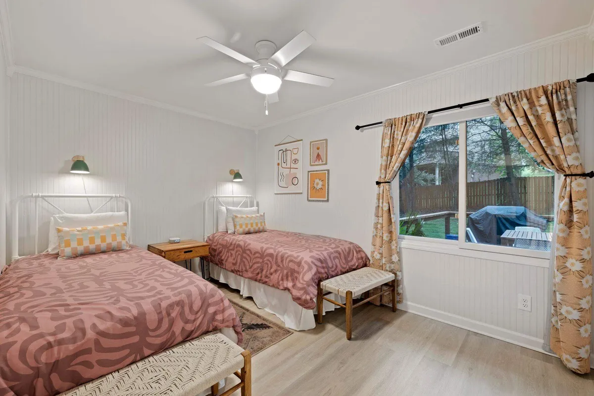 A bright bedroom with two twin beds, each adorned with patterned bedspreads, decorative wall art, and a large window letting in natural light. The space feels welcoming and playful, with ample floor space for relaxing or playing games after a day of adventure.