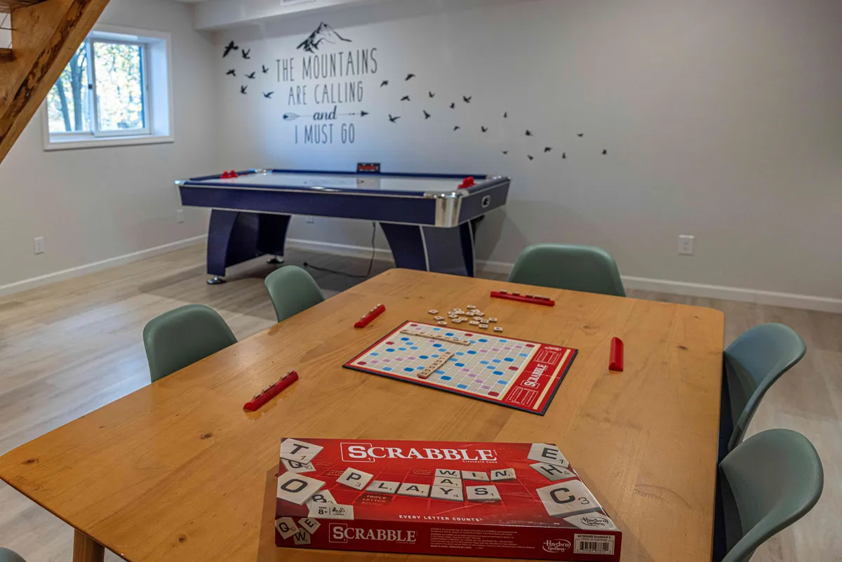 The image displays a vibrant game room with a Scrabble board set up on a table, an air hockey table nearby, and a wall mural featuring a mountain theme. The space feels bright, fun, and ready for group activities, including board games and air hockey.