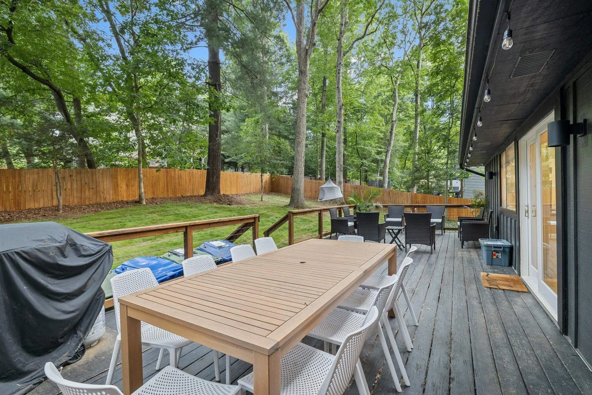 A spacious wooden deck is set with a large dining table surrounded by white outdoor chairs. The area is bordered by lush green trees and opens onto a fenced yard, creating an inviting atmosphere for gatherings and meals outside.