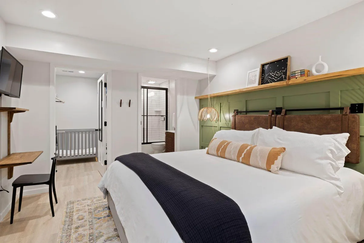 A serene bedroom featuring a king-sized bed with crisp white linens and decorative pillows, a green accent wall adorned with a wood shelf and subtle decor, a suspended light fixture, a TV mounted on the wall, and a small desk with chair beside the bed. Soft natural light filters through the window, enhancing the tranquil atmosphere.